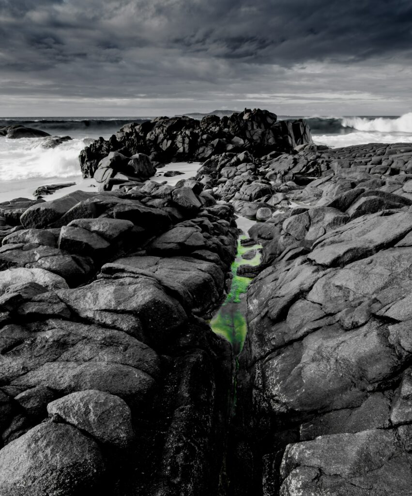 Dramatic black rocks on rugged coast with crashing waves and vibrant green tide pool background Wallpaper