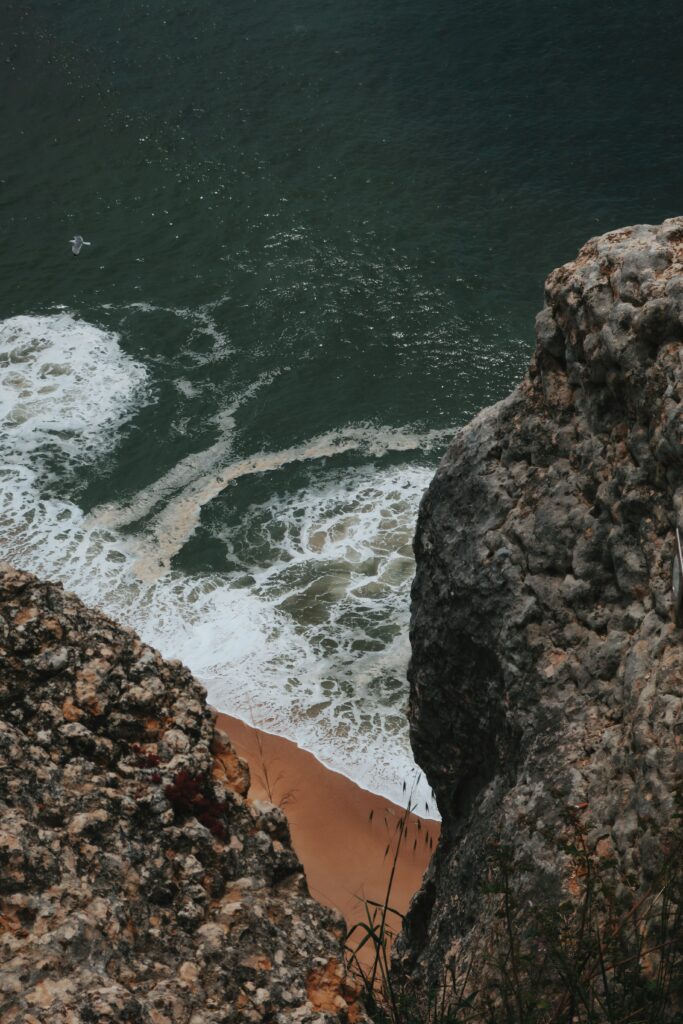 Dramatic seaside cliff with rugged rocks sandy beach and gentle waves on deep green sea wallpaper