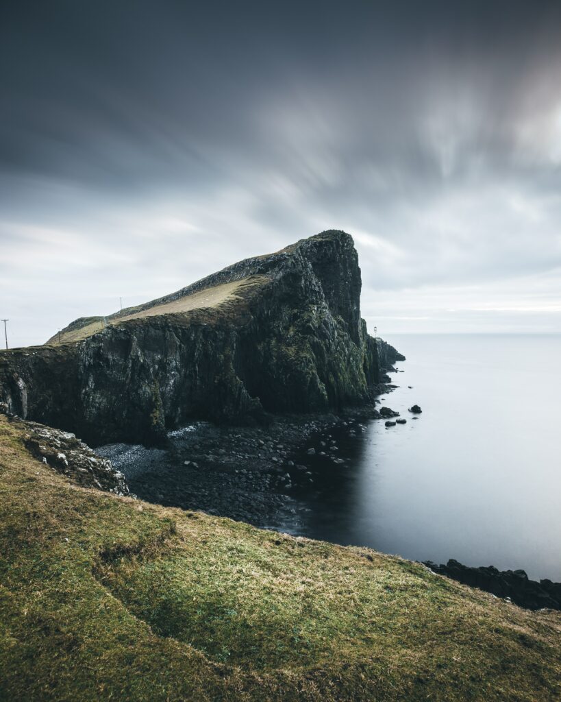 Towering coastal rock cliff with rugged stones and green grass overlooking calm sea under overcast sky Wallpaper
