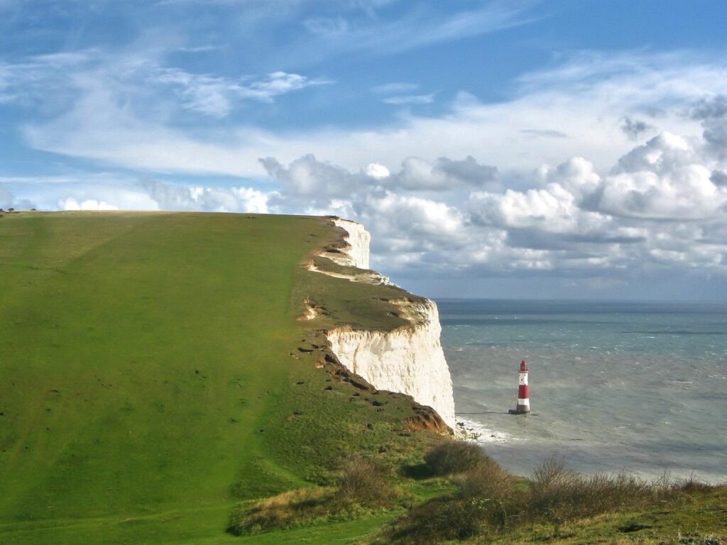 Dramatic white cliff with green grass and red white lighthouse by sea under cloudy sky wallpaper
