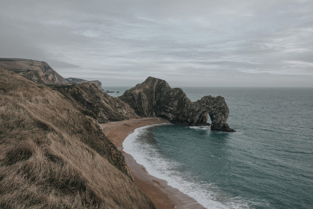 Dramatic coastal scene with rugged rocks waves white foam sandy shore and grassy cliffs under sky Wallpaper