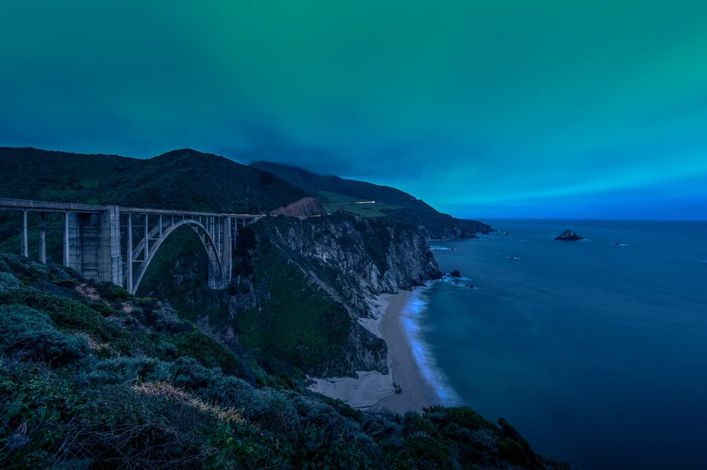 Dramatic coastal dusk with rugged rocks sandy beach arched bridge glowing sea blue green sky Wallpaper