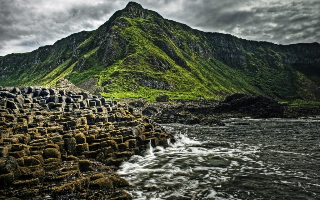 Dramatic coastal wallpaper with hexagonal stones waves lush green mountains and moody sky