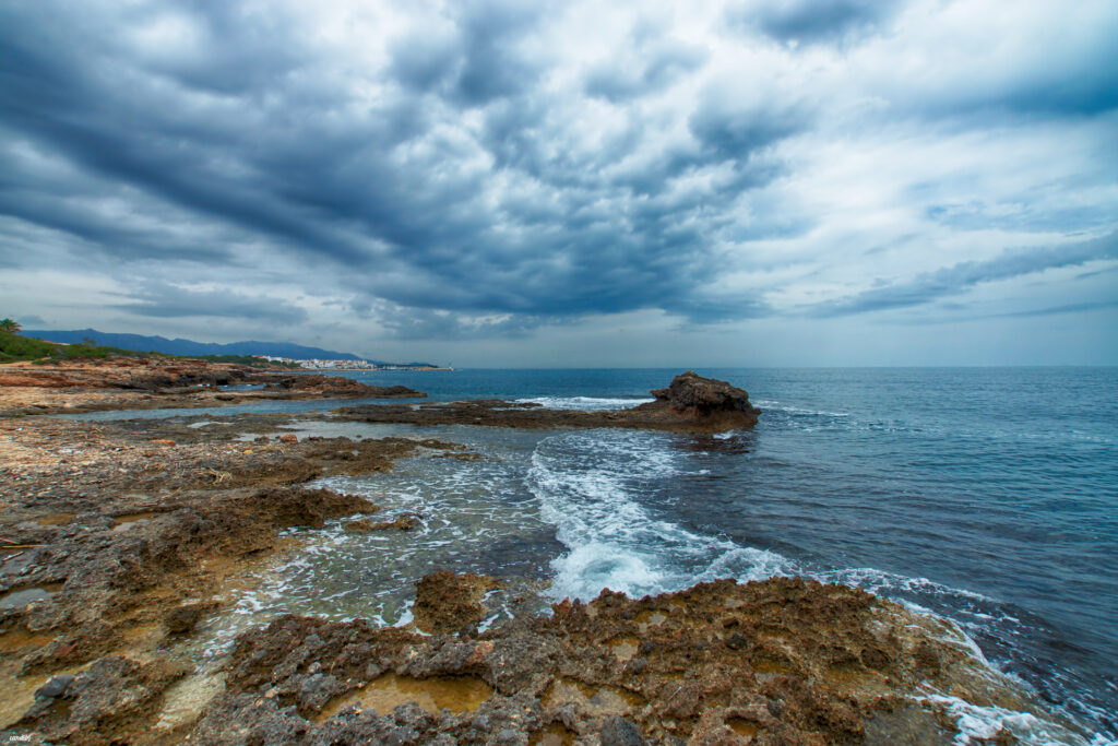 Dramatic coastal landscape with rugged rocks gentle waves moody clouds vivid blue earthy tones Wallpaper