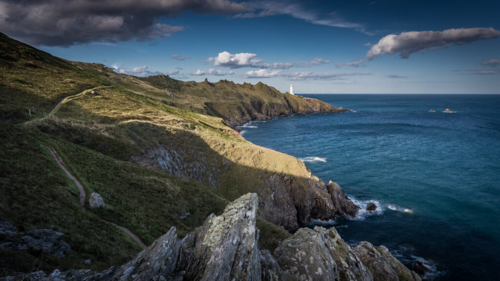 Rugged coastal rocks with blue sea and white lighthouse under dramatic sunlight and clouds Wallpaper