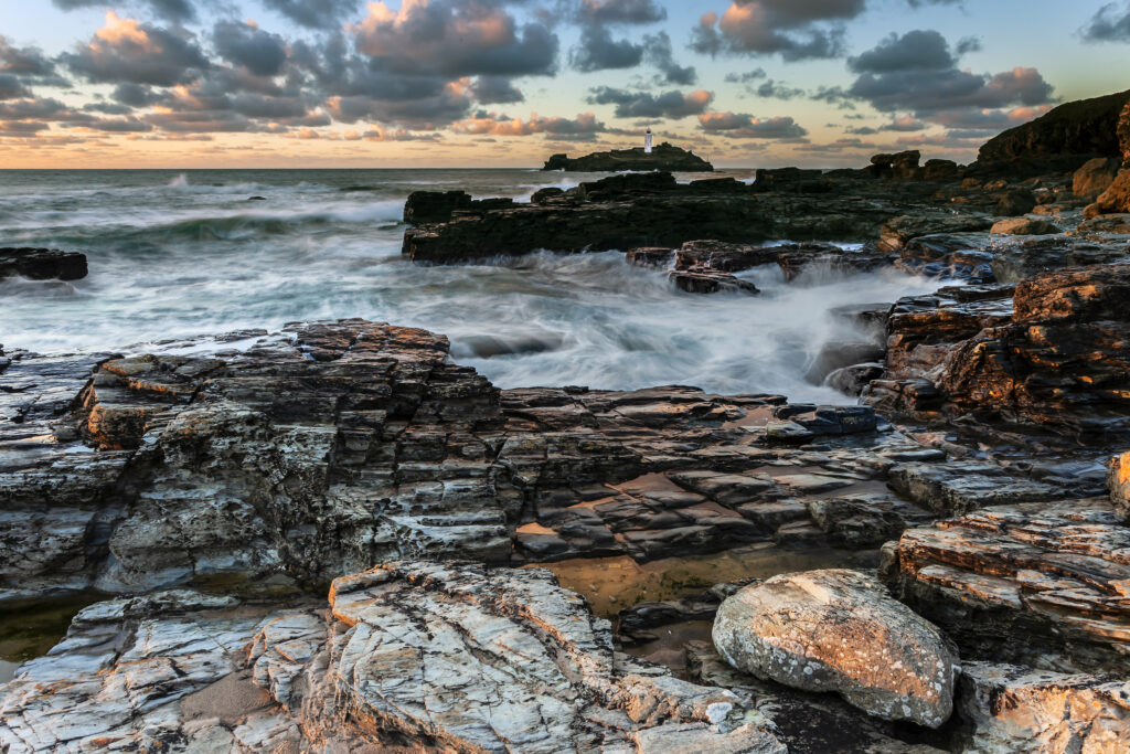 Rugged coastal landscape with crashing waves lighthouse on rocky island under vibrant sky wallpaper