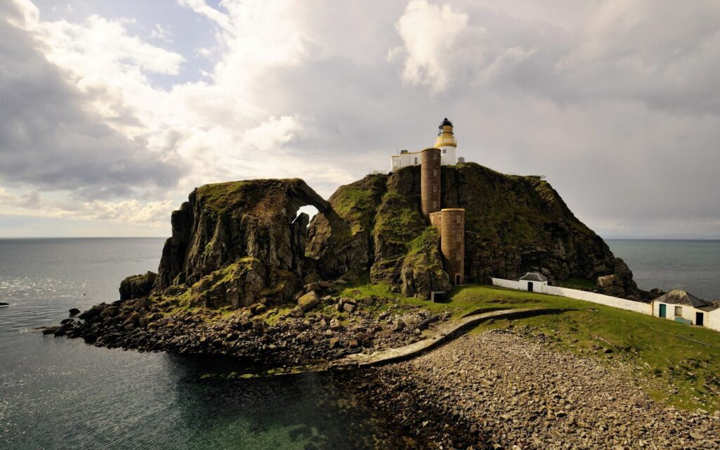 Historic lighthouse on rocky coastal cliffs with sunlight highlighting weathered stone and ocean Wallpaper