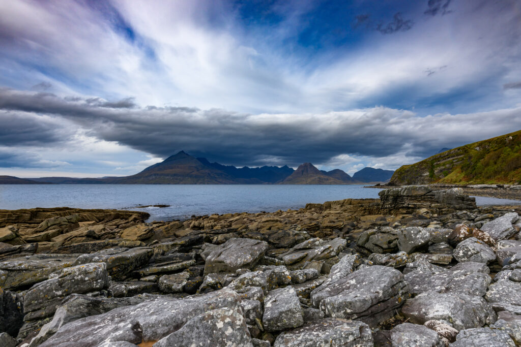 Dramatic coastal landscape with rugged rocks serene sea and towering mountains under moody sky Wallpaper