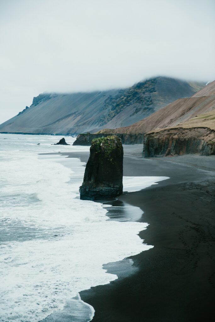 Dramatic coastal rock formation on black sand beach with ocean waves and misty mountains background Wallpaper