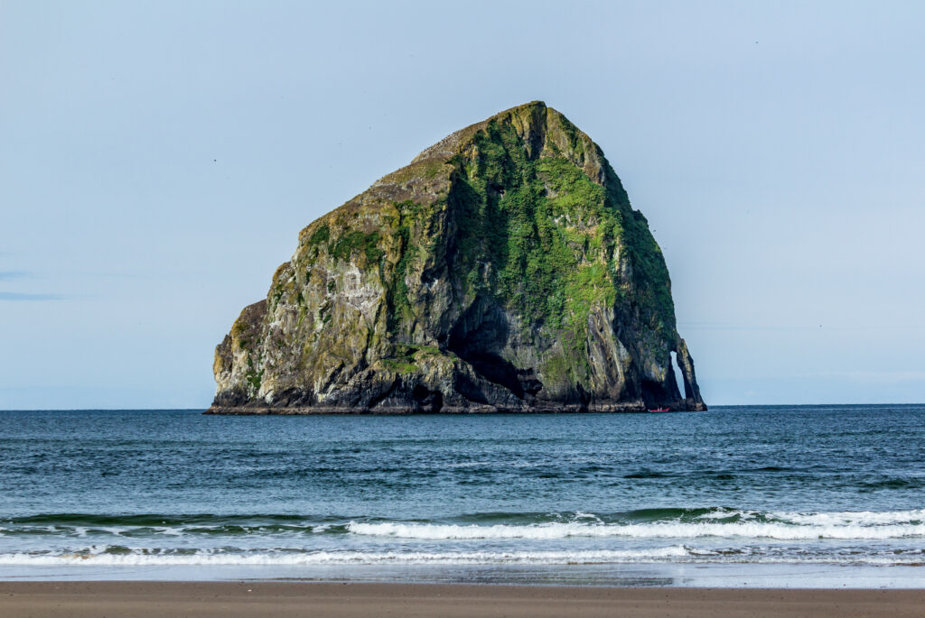 Massive coastal rock formation with green vegetation rising from calm blue ocean under clear sky Wallpaper