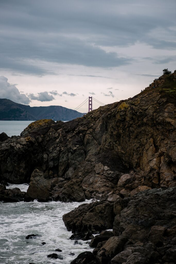 Dramatic coastal rocks with crashing waves and Golden Gate Bridge under overcast sky wallpaper