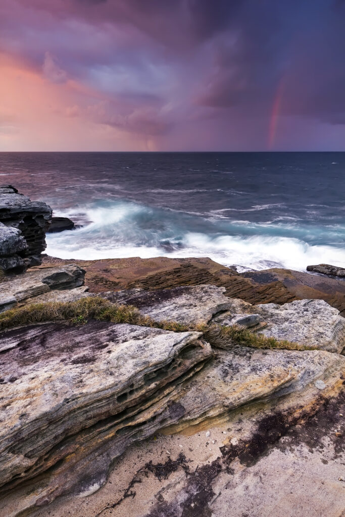 Rugged coastal rocks with crashing waves under vibrant sunset sky and faint rainbow horizon wallpaper