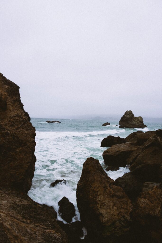 Dramatic coastal scene with jagged rocks and crashing waves under misty cloudy sky wallpaper background