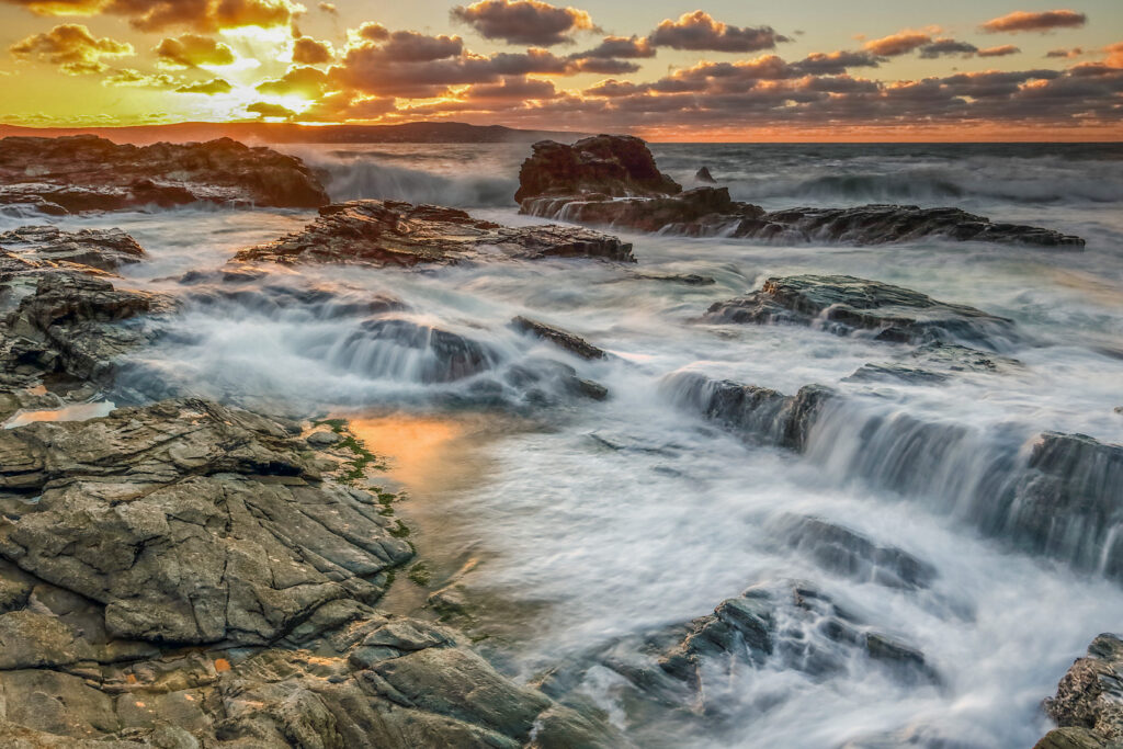 Rugged rocky coastline with crashing waves under a vivid golden blue sunset seascape wallpaper