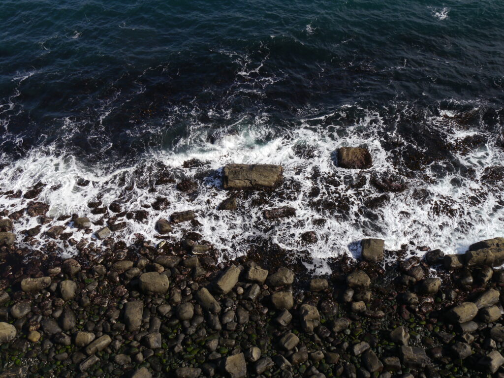 Aerial view of rugged coastal shore with pebbles foamy waves and deep blue ocean water wallpaper