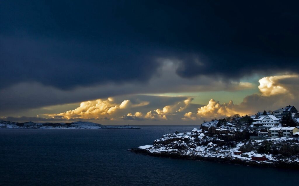 Dramatic coastal scene with snowy rocky shore charming houses deep blue sea and stormy sky wallpaper