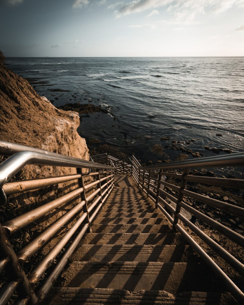 Dramatic staircase descending rocky cliffs to calm sea with warm sunlight and coastal scenery Wallpaper