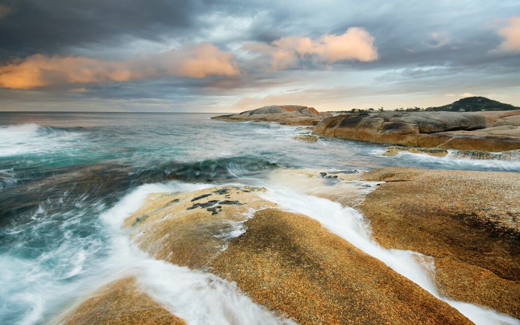 Dramatic coastal scene with turquoise waves over sandy rocks and golden sunset sky wallpaper background