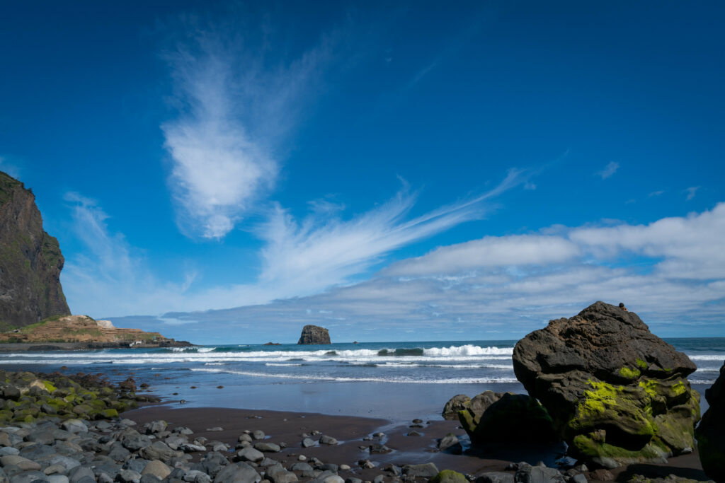Dramatic seashore with rugged rocks smooth pebbles black sand waves crashing vivid blue sky Wallpaper