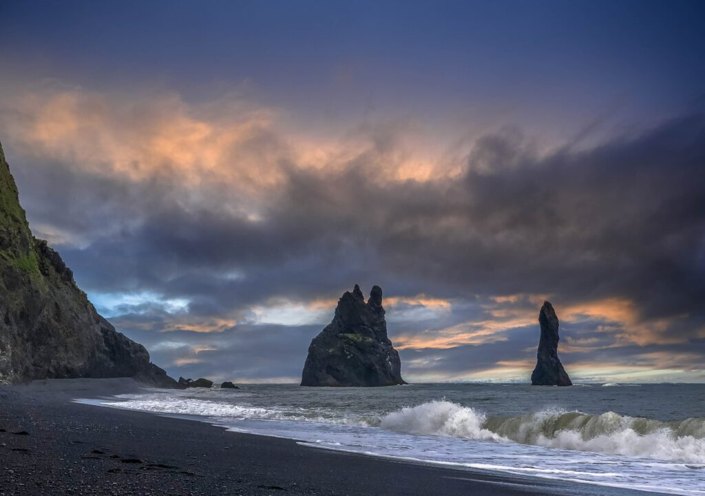 Dramatic coastal wallpaper with rugged rocks waves crashing on black sand beach at dusk sky