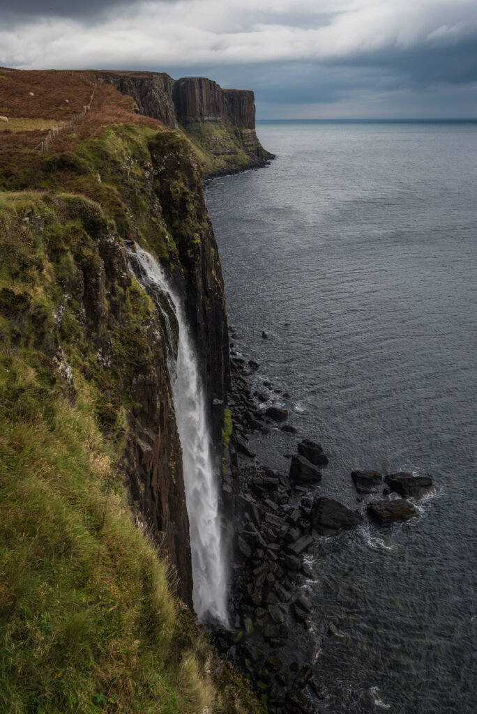 Tall waterfall cascades rocky cliffs into calm sea with green grass under moody overcast sky Wallpaper