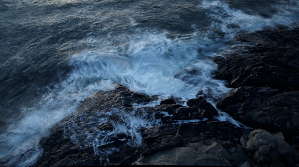 Dynamic ocean waves crashing on rugged dark stones with white foam in a serene coastal scene Wallpaper