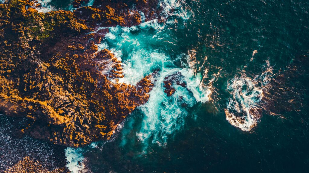 Aerial view of rugged rocks meeting deep blue sea with crashing waves and white foam coastline Wallpaper