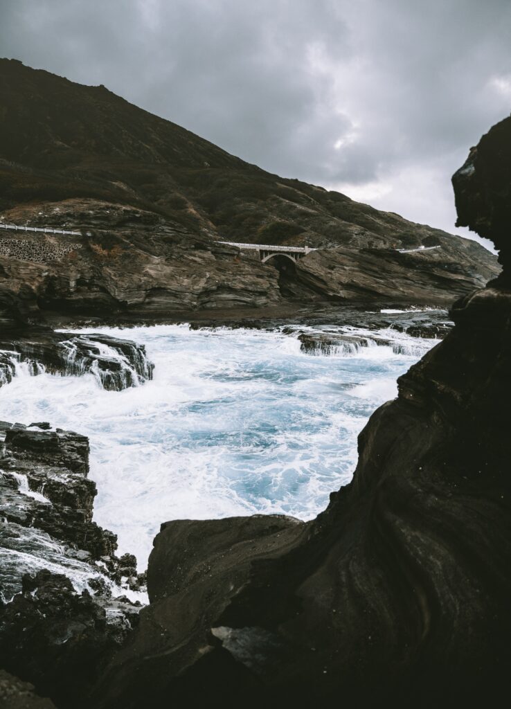 Dramatic coastal waves crash on rugged rocks beneath moody sky with narrow bridge in background Wallpaper