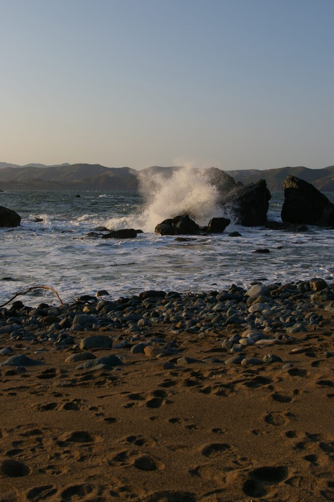 Dramatic crashing waves on rugged stones with smooth pebbles sandy footprints coastal shore Wallpaper