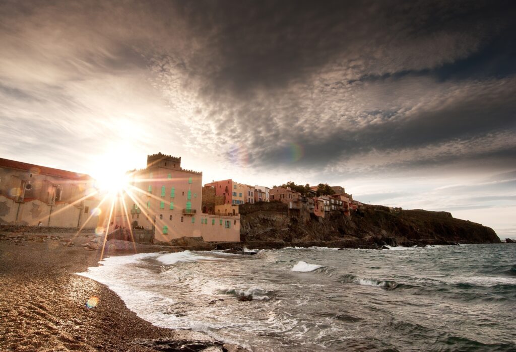 Dramatic coastal waves on pebbled shore sunlight on cliff homes under swirling cloud sky wallpaper