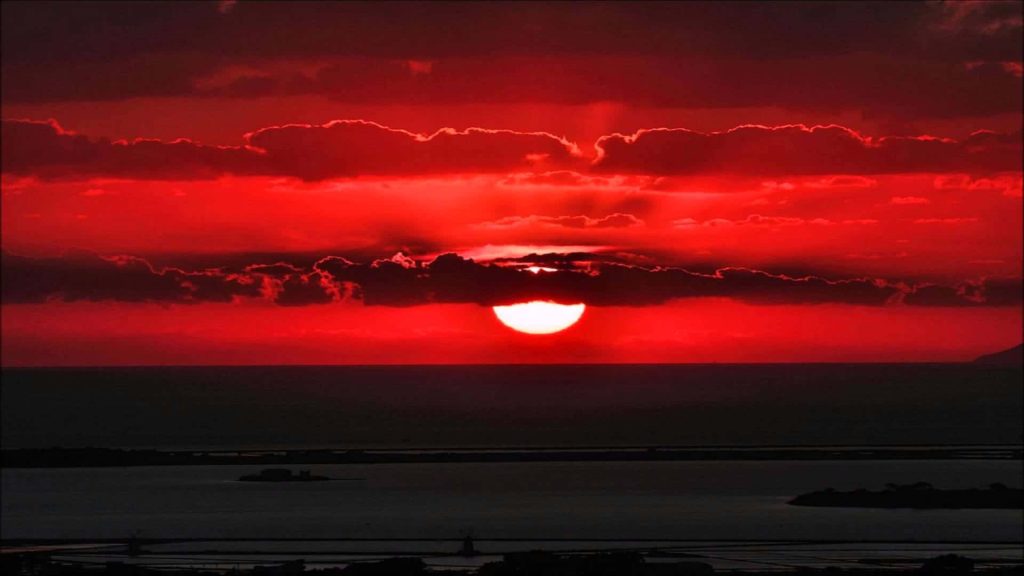 Glowing sun setting behind dark clouds casting vibrant crimson hues over calm waters sunset wallpaper
