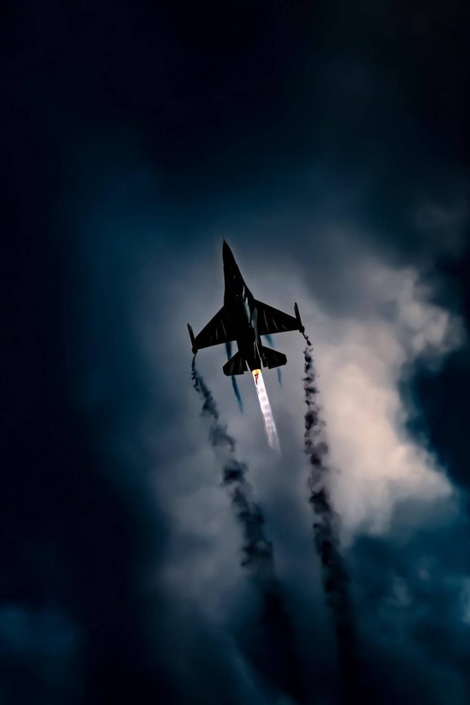 Silhouetted F-16 fighter jet soaring with afterburner and smoke trails on dark cloudy sky background Wallpaper