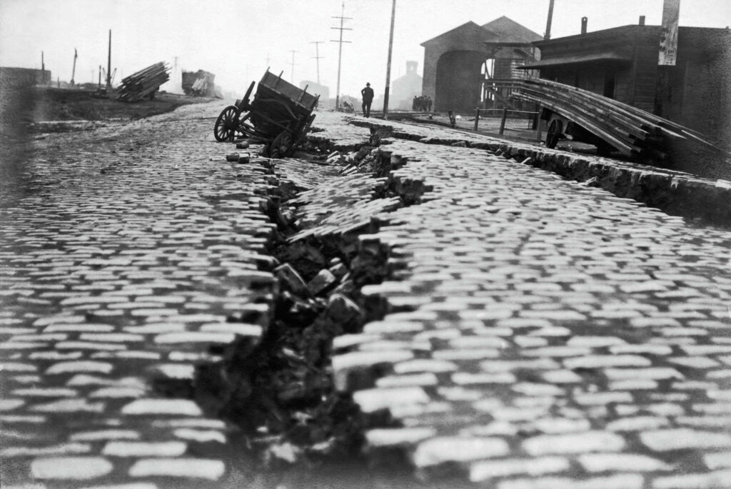 Monochrome earthquake aftermath with cracked cobblestone street toppled cart and lone figure Wallpaper