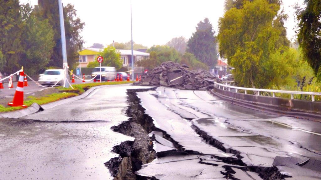 Cracked uplifted road after earthquake with debris under cloudy sky and blurred traffic background Wallpaper