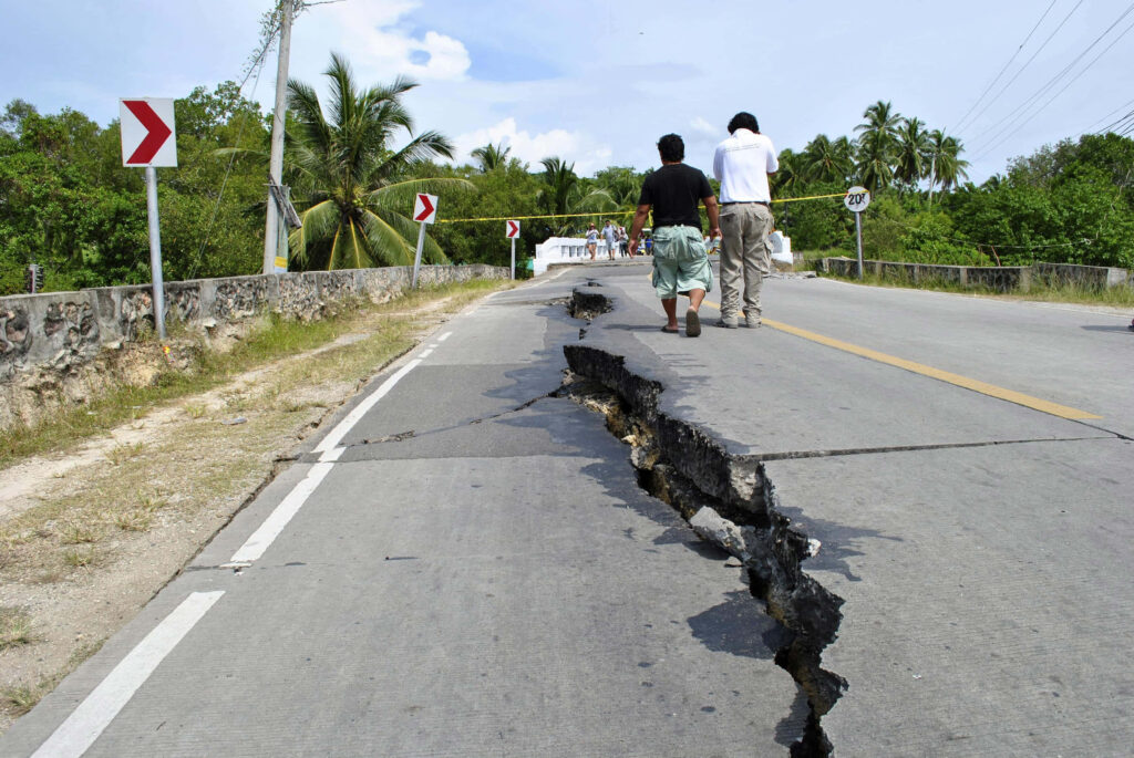 Earthquake damage with cracked road lush trees red signs two people walking under clear sky Wallpaper