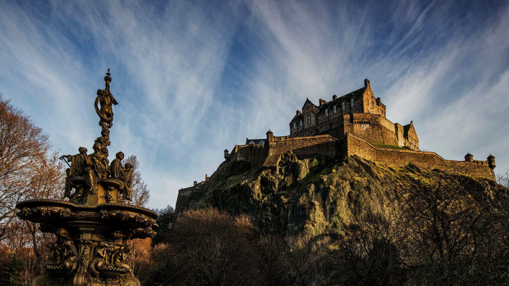Edinburgh Castle on rugged cliffs under dramatic clouds with ornate fountain in warm golden light Wallpaper
