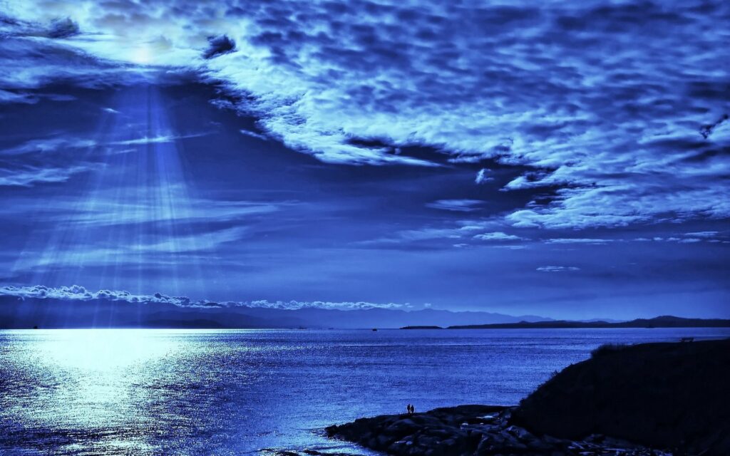 Beams of light pierce dark blue evening clouds over calm sea with silhouettes on rocky shore Wallpaper