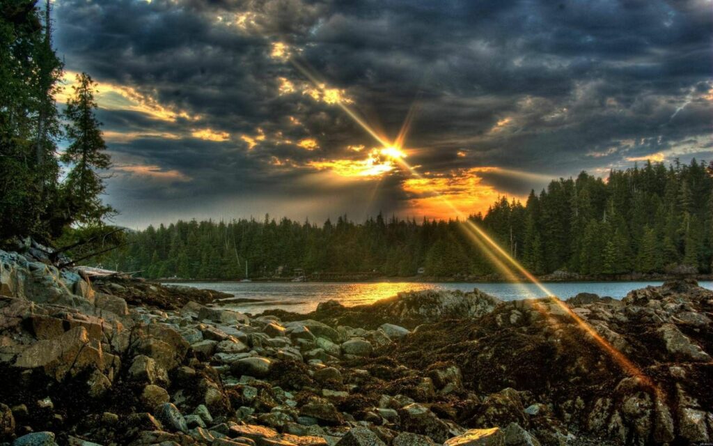 Dramatic sunset over tranquil lake with sunbeams dark clouds rocky shore and dense forest Wallpaper