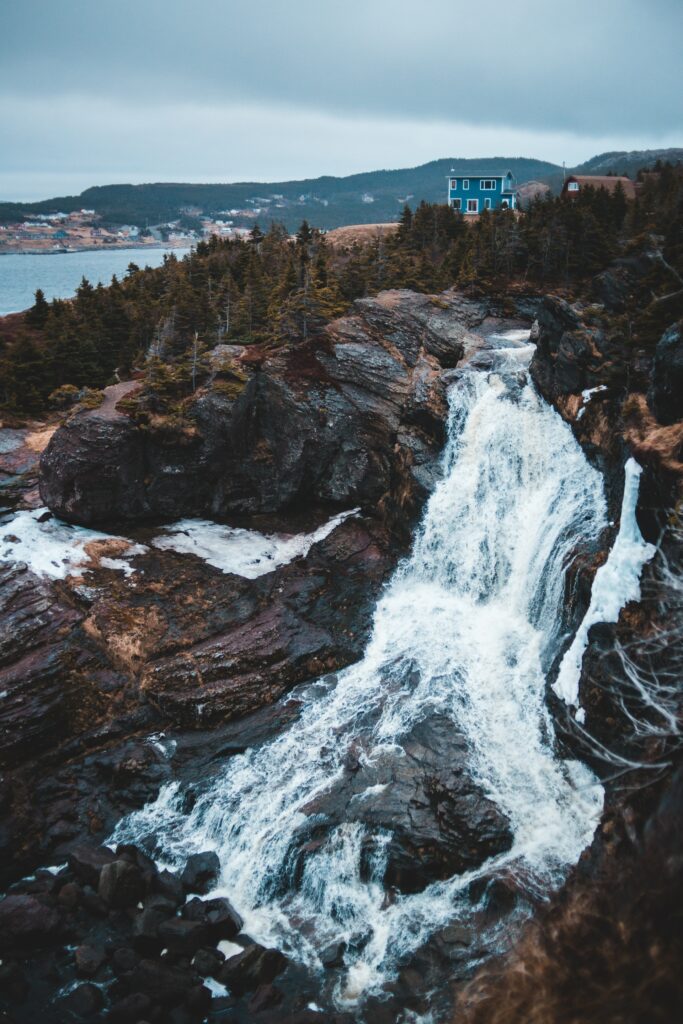 Foamy fast river cascades over rocky shore near cliffs dense trees with house and moody sky wallpaper