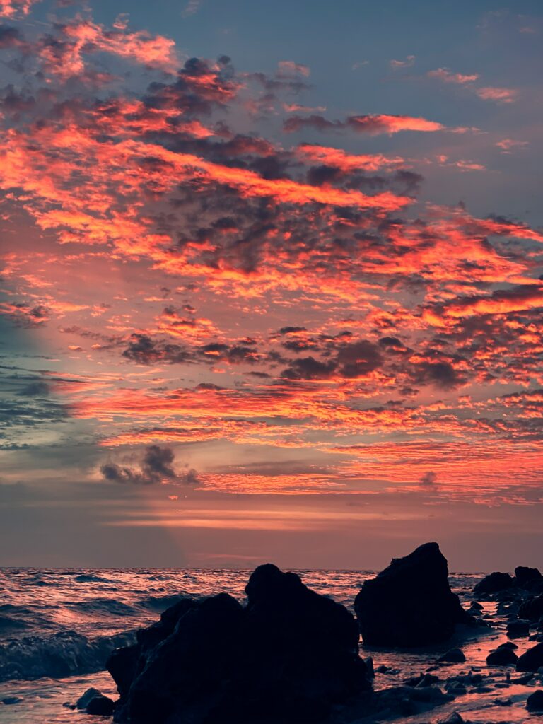 Fiery sunset sky with orange pink clouds over dark seaside rocks and gentle rolling waves Wallpaper