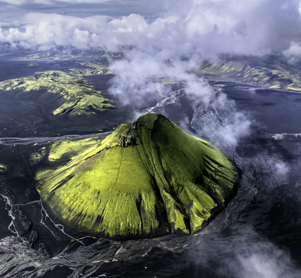Aerial view of vibrant green Icelandic volcano with cloud-covered peak and surrounding rivers Wallpaper