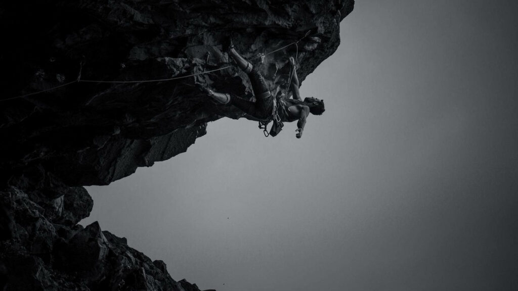Greyscale image of a focused man rock climbing a steep cliff with a minimalist background wallpaper