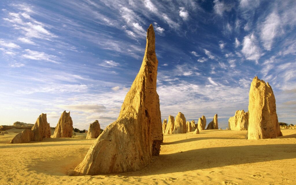Jagged sandstone formations rise from golden desert sands under vivid blue sky with wispy clouds Wallpaper