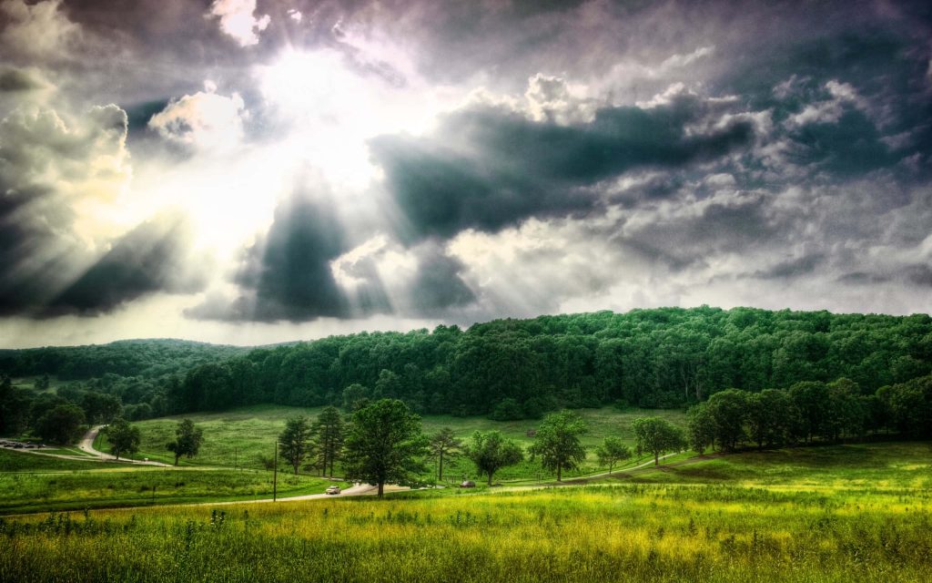 Dramatic landscape with green meadows winding road and sunbeams piercing moody cloud sky wallpaper