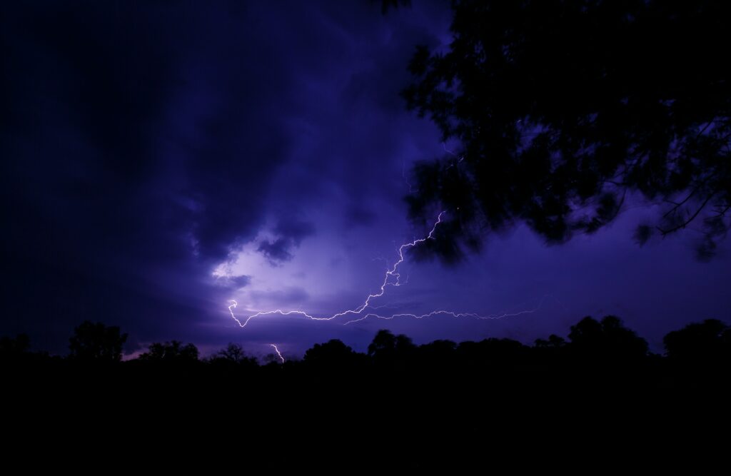 Dramatic lightning bolt streaks dark stormy sky illuminating purple clouds and silhouetted trees Wallpaper