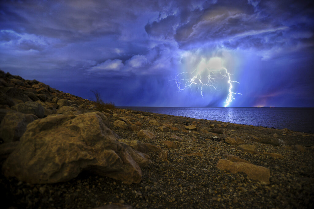Lightning bolt lights overcast sky above calm lake and rugged rocks during stormy night wallpaper