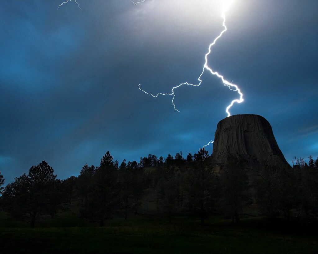Lightning bolt strikes above towering rock formation at night with dark woods and silhouetted trees Wallpaper