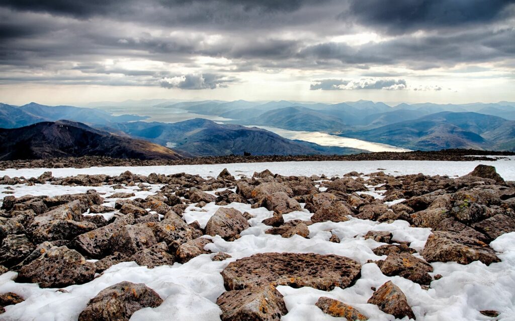 Rugged rocks on melting snow with distant mountains and cloudy sky nature landscape wallpaper