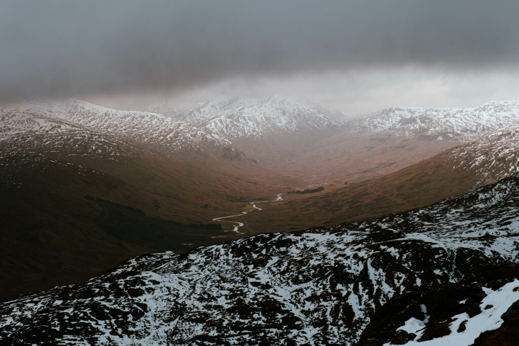 Snowy mountain peaks with winding river and rugged brown terrain under moody sky wallpaper