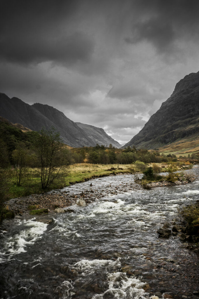 Dramatic mountain landscape with rugged rocks flowing stream and lush trees under cloudy sky Wallpaper